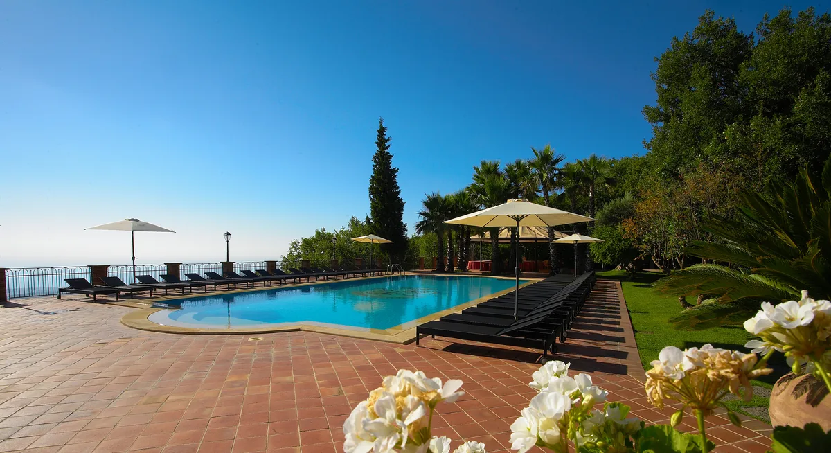Piscina esterna con lettini e ombrelloni circondata da palme e vista sul mare