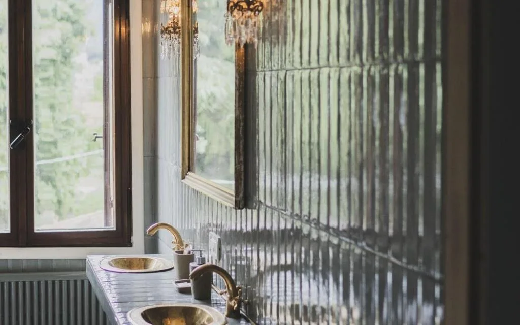 Bagno con doppio lavabo in ottone, piastrelle lucide e finestra affacciata sul verde