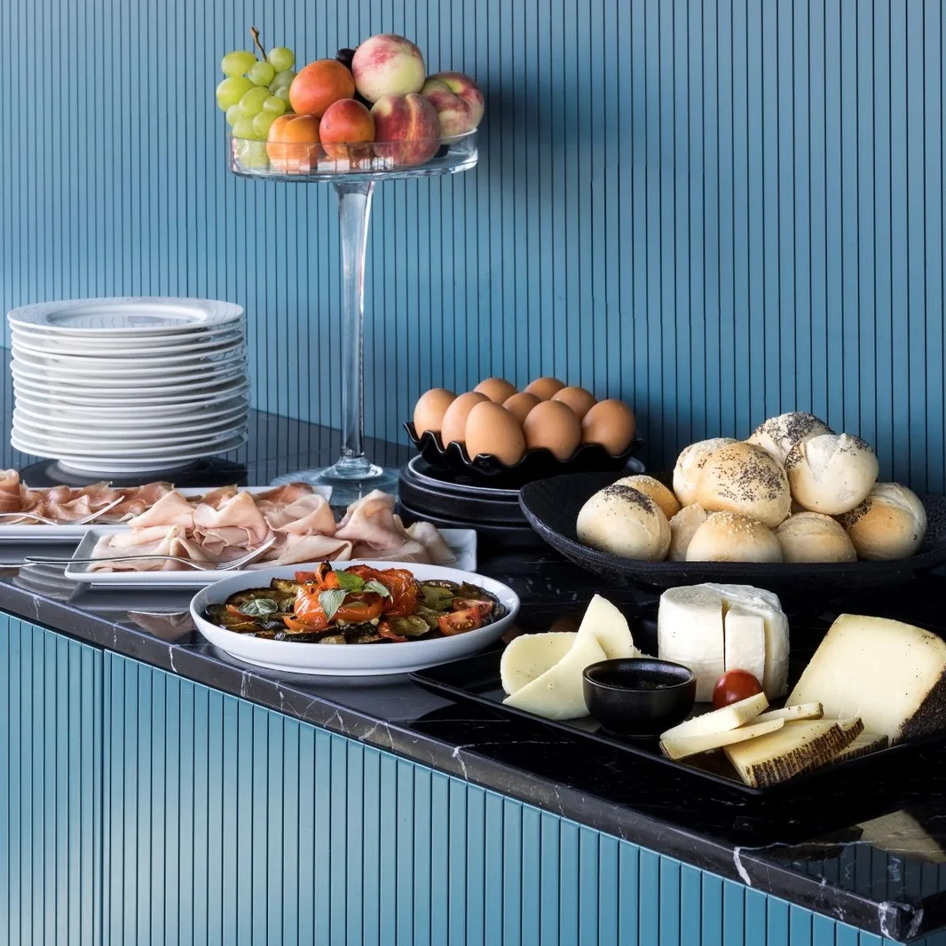 Buffet della colazione con salumi, formaggi, uova, pane e frutta fresca su parete blu