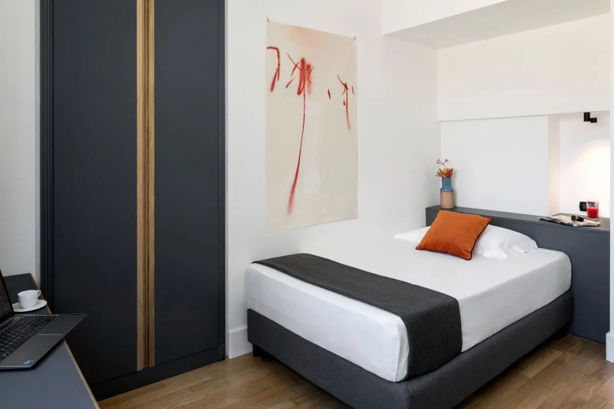 Camera singola con letto, cuscino arancione, armadio scuro e zona lavoro
