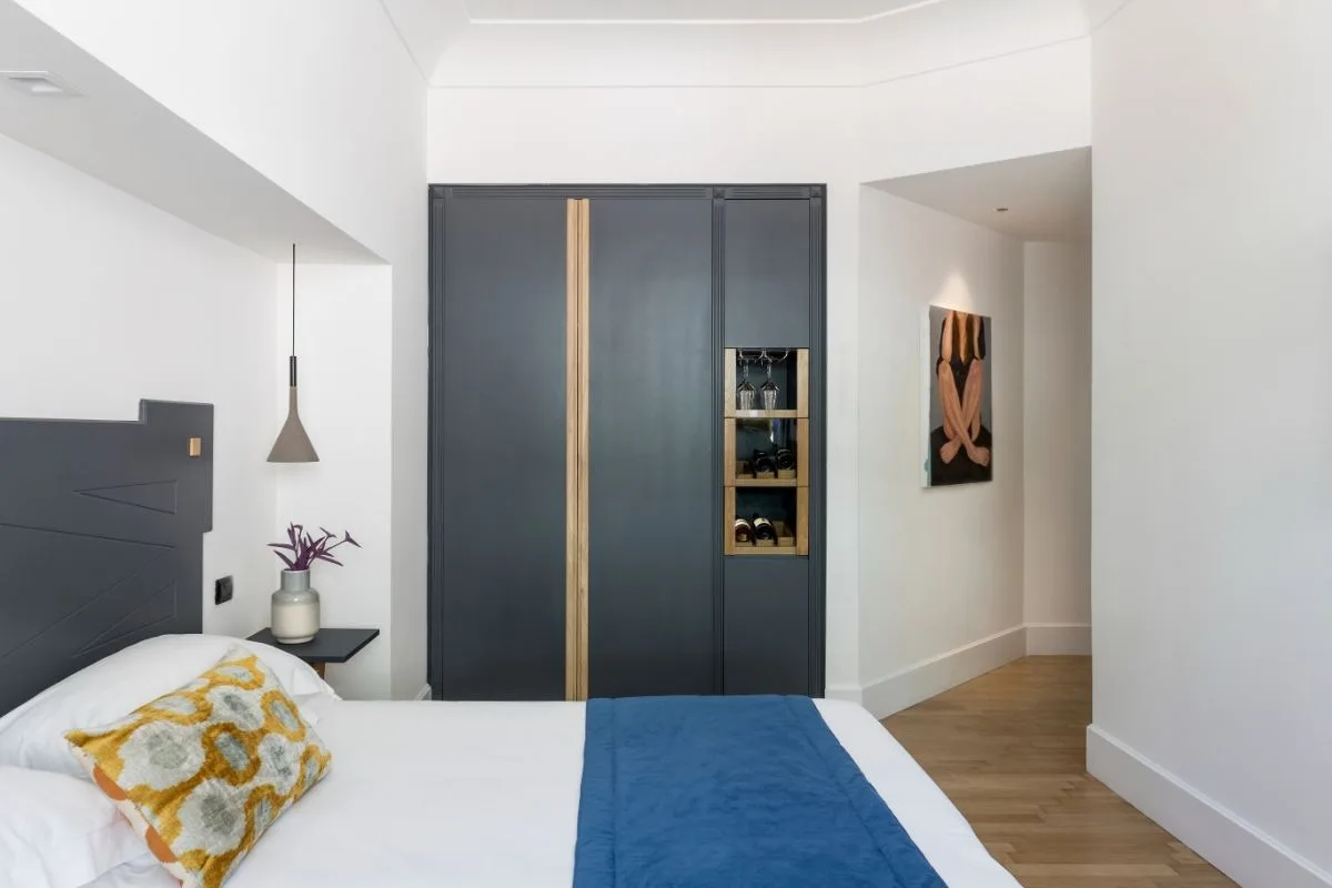 Camera con testiera grigia, armadio scuro con cantinetta vini e opera d'arte lungo il corridoio