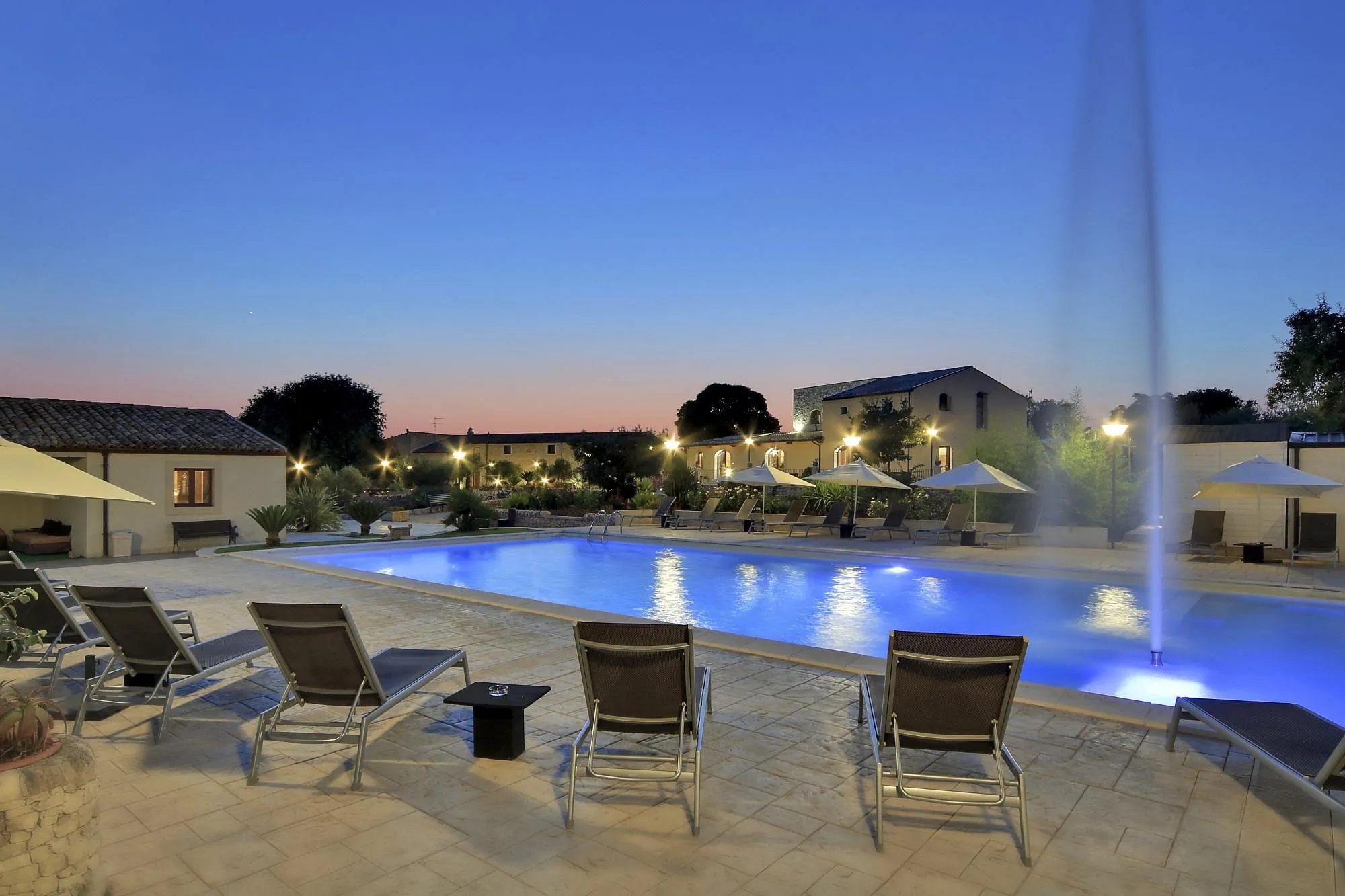 Piscina illuminata al crepuscolo con lettini, ombrelloni e getto d'acqua