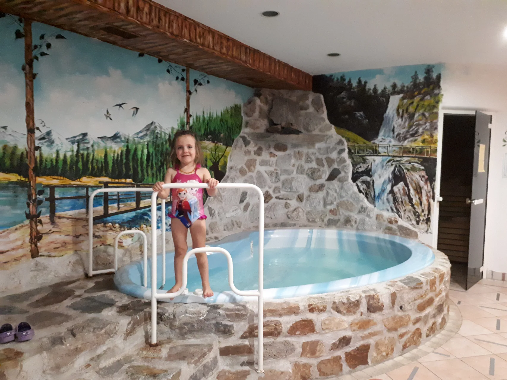 Piccola piscina interna con bambina, pareti decorate con murales di montagne e cascate