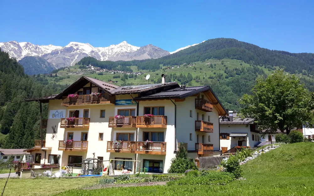 Facciata dell'Hotel Ortles con balconi fioriti e vista sulle montagne innevate in estate