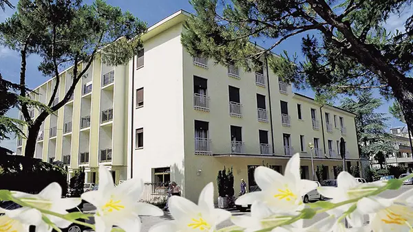 Facciata esterna dell'hotel con giardino, alberi e fiori bianchi in primo piano