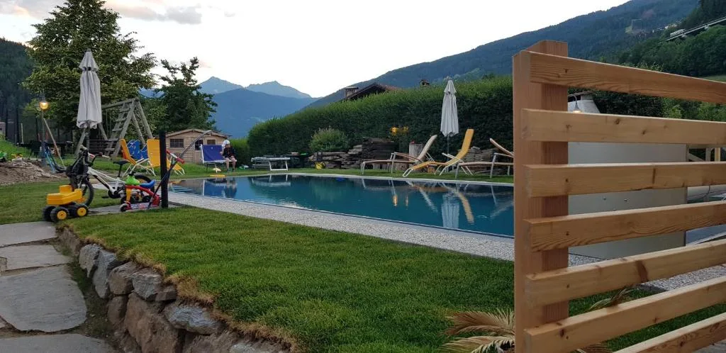Piscina esterna con area giochi per bambini, ombrelloni e lettini circondata da verde e montagne