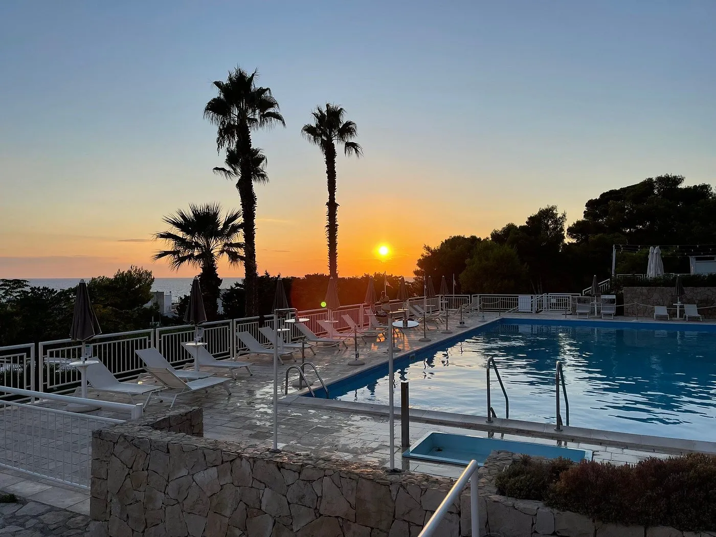 Piscina al tramonto con palme e sole che cala sul mare