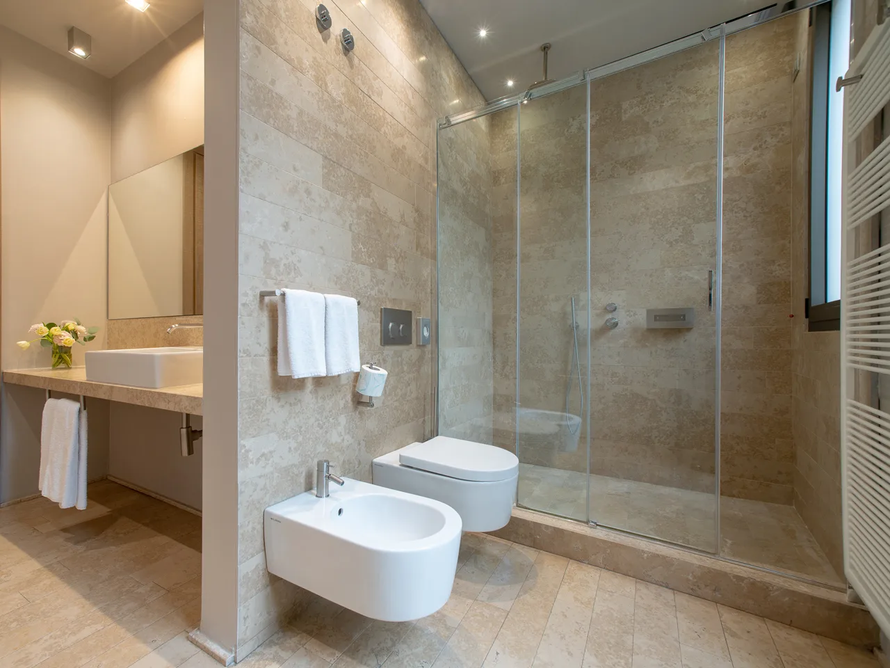 Bagno moderno in pietra beige con doccia in vetro, bidet, WC e lavabo con specchio