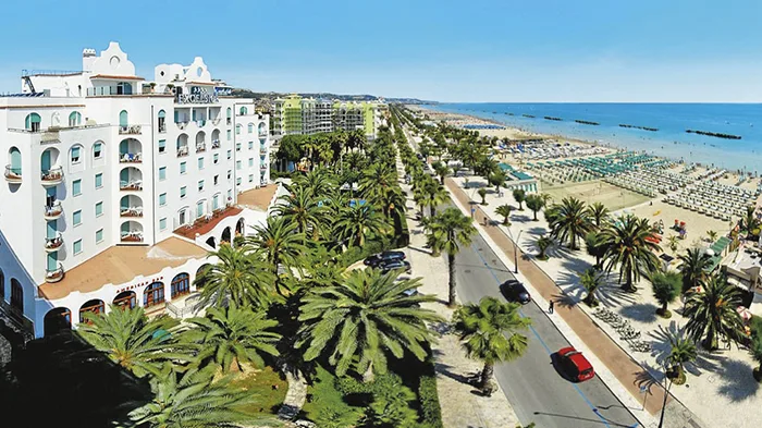 Veduta panoramica dell'hotel bianco sul lungomare con palme, spiaggia attrezzata e mare