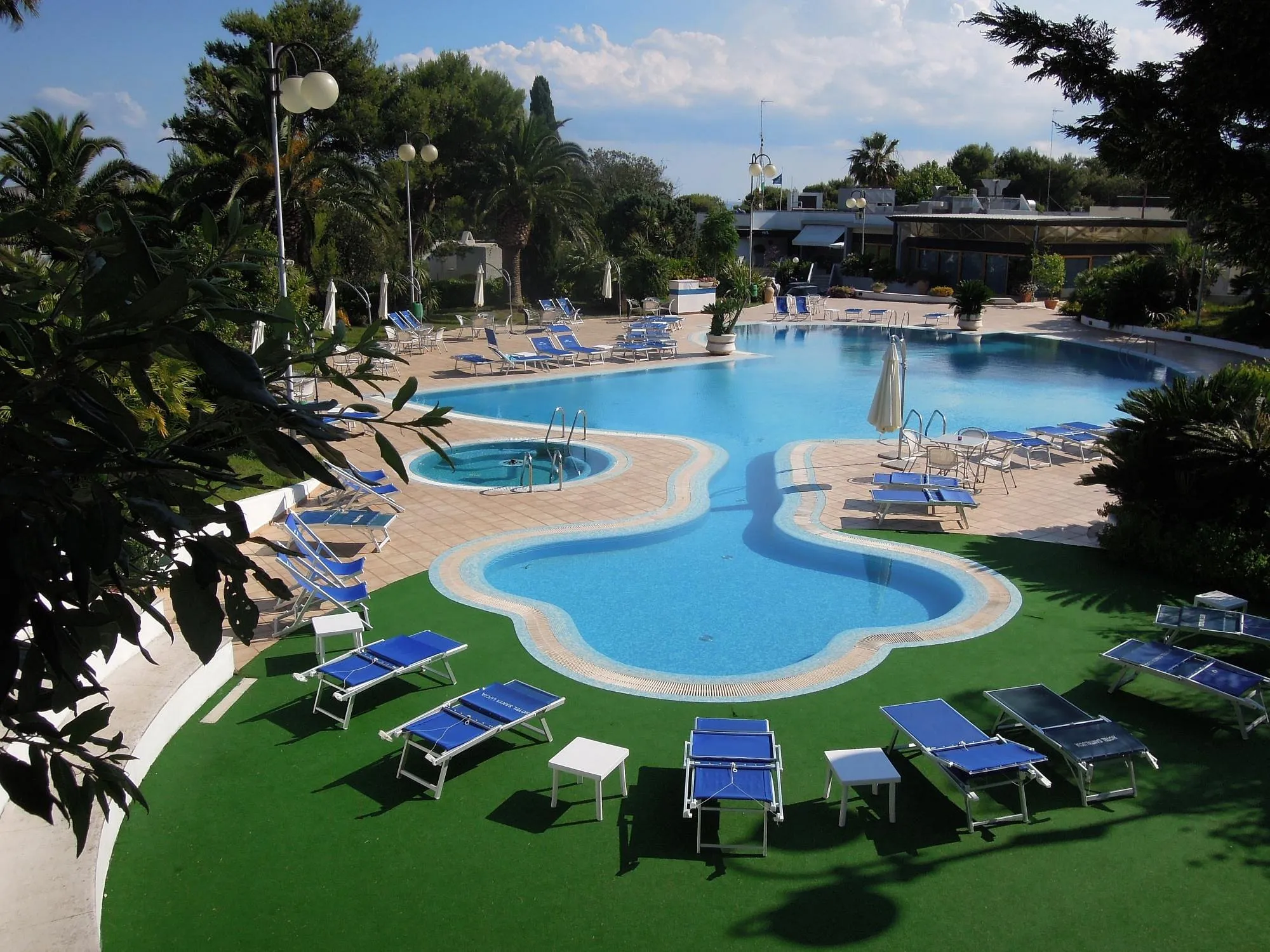 Piscina panoramica con prato verde, lettini blu e ombrelloni immersa in un giardino di palme
