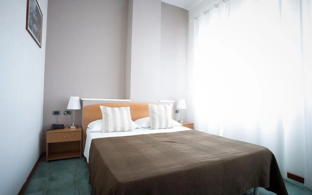 Camera doppia con letto matrimoniale, comodini in legno e ampia finestra con tende bianche
