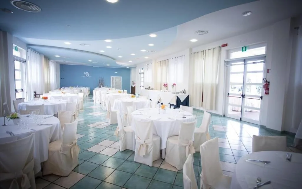Sala ristorante con tavoli rotondi apparecchiati, sedie con coprisedia bianchi e soffitto azzurro