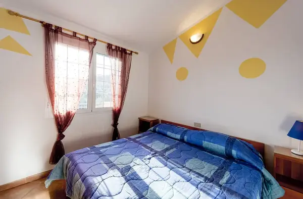 Camera da letto con pareti decorate gialle, finestra con tende e letto matrimoniale blu