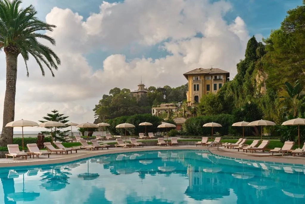Piscina con lettini e ombrelloni circondata da palme e giardini, con villa storica sullo sfondo