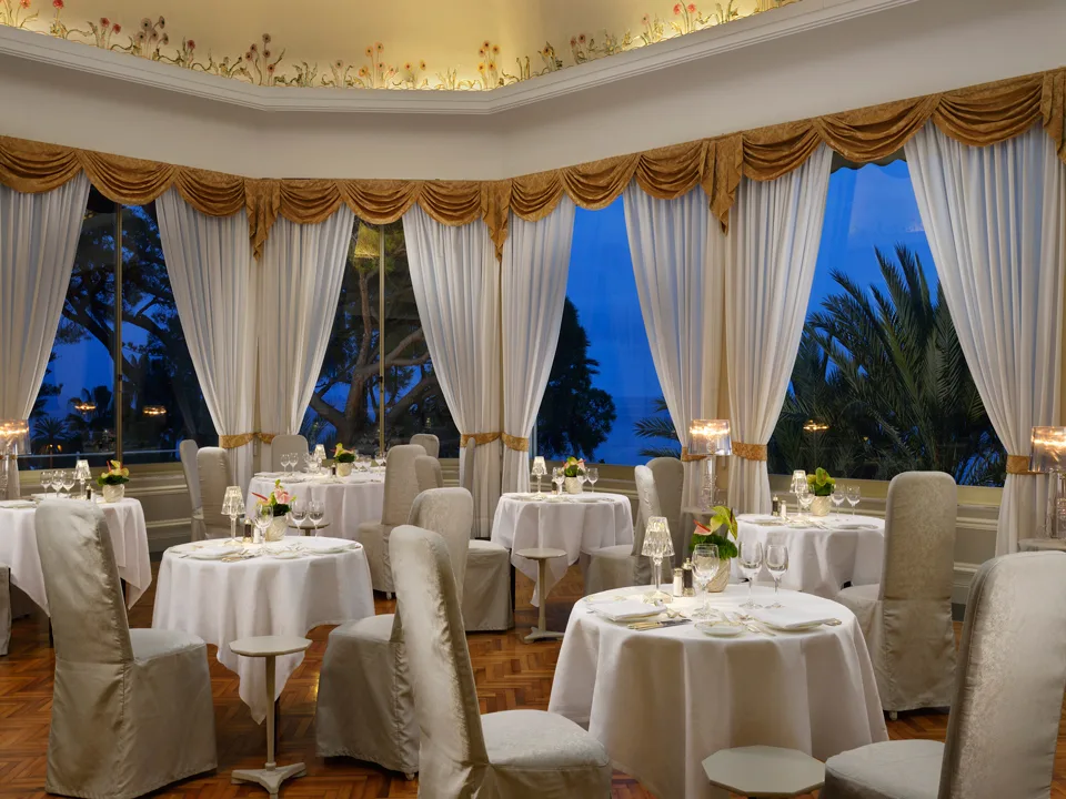 Elegante sala ristorante interna con tavoli rotondi, tendaggi dorati e vista serale sui giardini