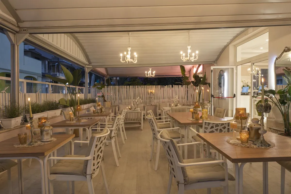 Sala da pranzo serale con tavoli in legno, sedie bianche e candele dorate
