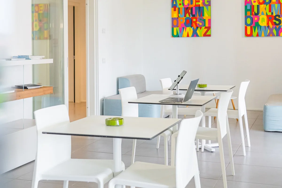 Sala comune luminosa con tavoli bianchi, laptop e quadri colorati con lettere