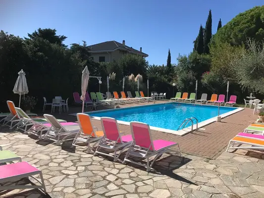 Piscina esterna con lettini colorati e ombrelloni, circondata da vegetazione mediterranea e cipressi