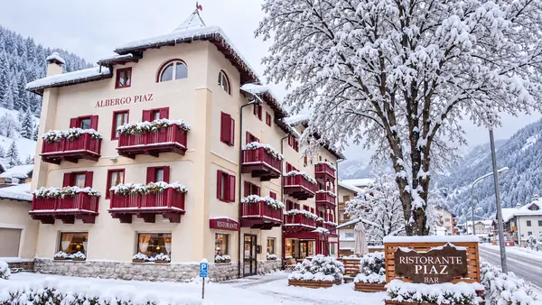 Facciata dell'Albergo Piaz in inverno con balconi rossi fioriti e grande albero innevato