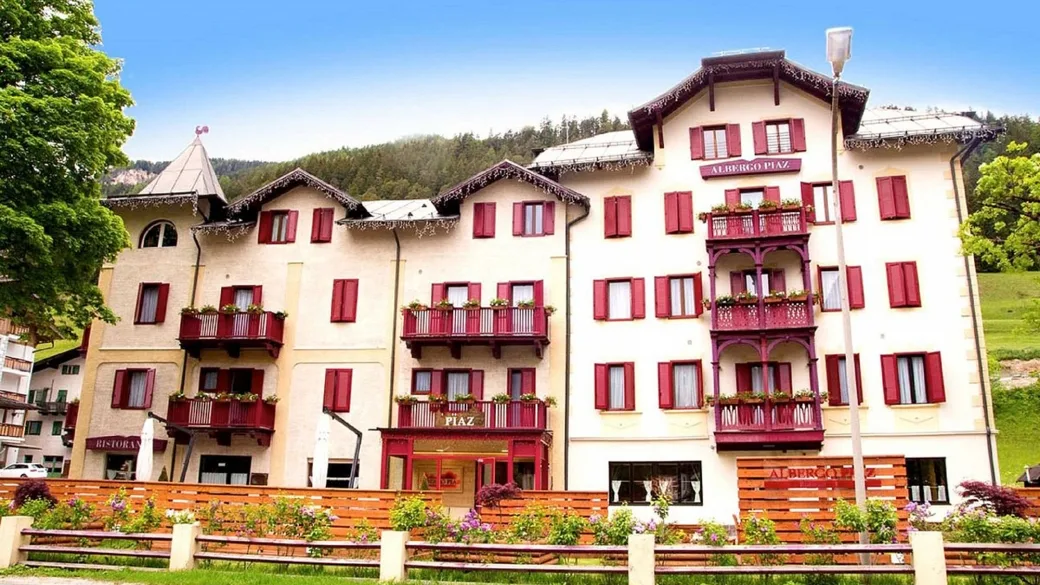Facciata dell'Albergo Piaz con persiane rosse, balconi fioriti e prato verde in estate