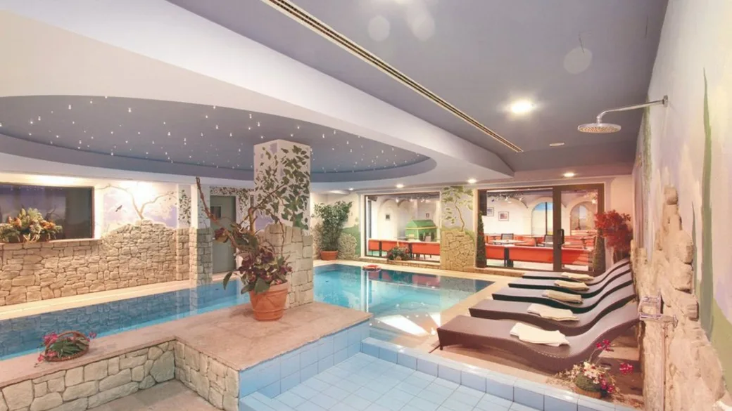 Piscina interna con lettini moderni, colonna decorata e pareti in pietra con murales