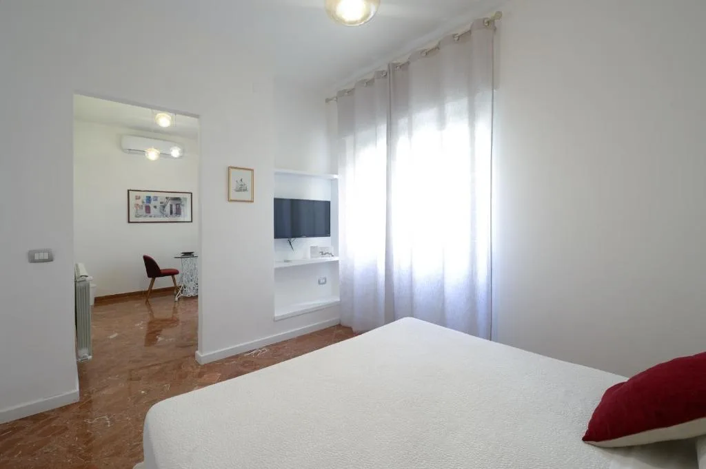 Camera da letto bianca con letto matrimoniale, TV a parete e vista su area soggiorno