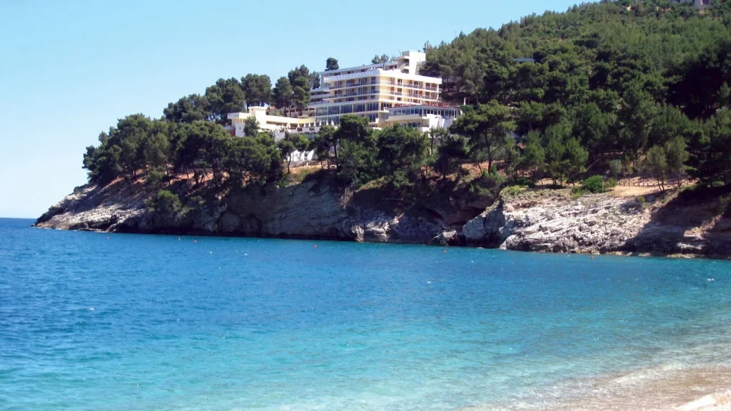 Hotel bianco su promontorio roccioso ricoperto di pini affacciato sul mare limpido