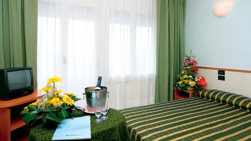 Camera con letto matrimoniale, tende verdi, televisore e tavolino con fiori e spumante