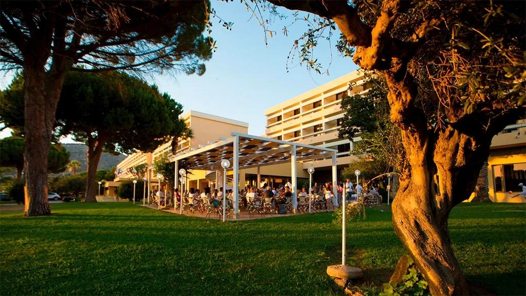 Esterno dell'hotel al tramonto con prato verde, alberi e terrazza affollata
