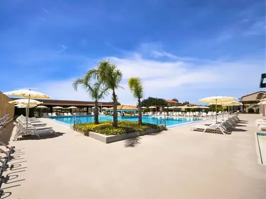 Piscina con palme centrali, ombrelloni gialli e ampio solarium sotto il cielo azzurro