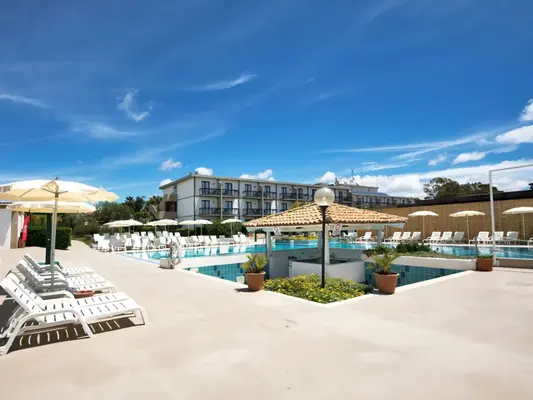 Piscina con lettini bianchi, ombrelloni e edificio dell'hotel sullo sfondo sotto cielo azzurro
