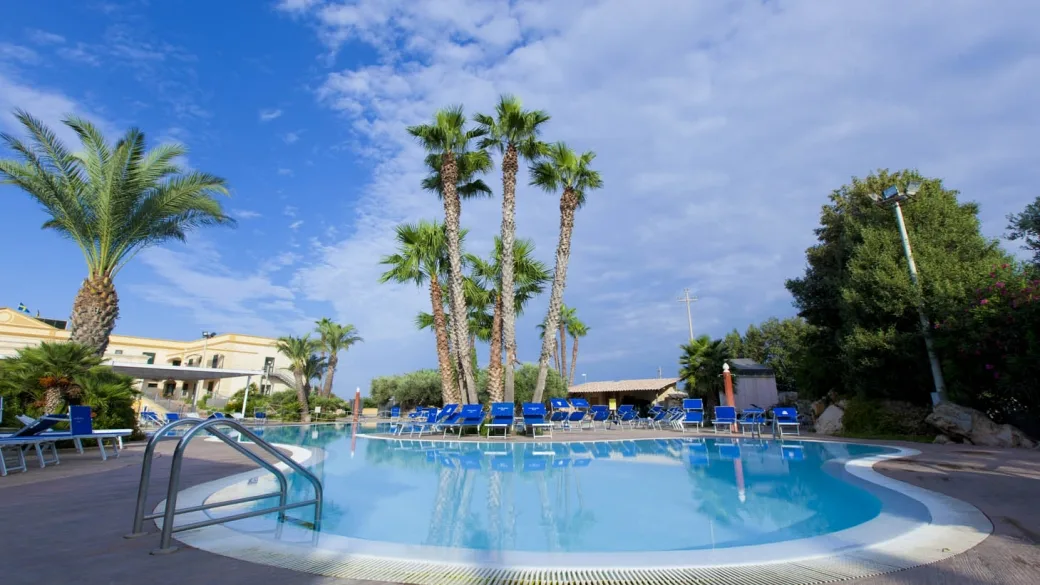 Piscina con palme svettanti, lettini e ombrelloni blu sotto il cielo azzurro