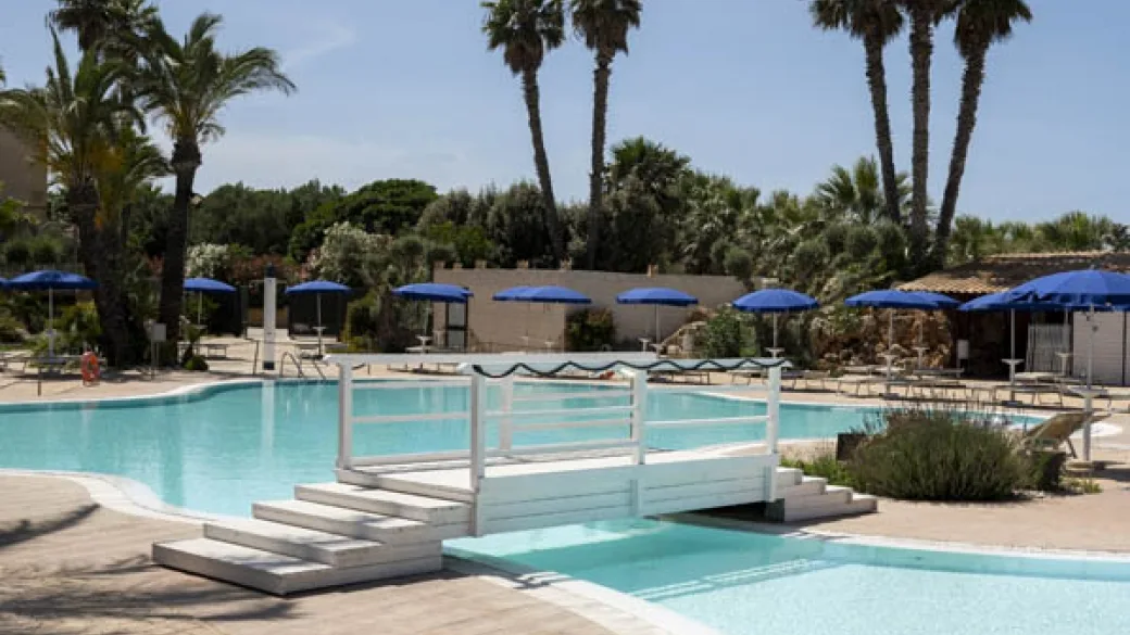 Piscina con ponticello bianco, ombrelloni blu e vegetazione mediterranea