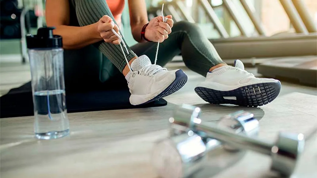 Donna allaccia le scarpe da ginnastica in palestra accanto a borraccia e manubri