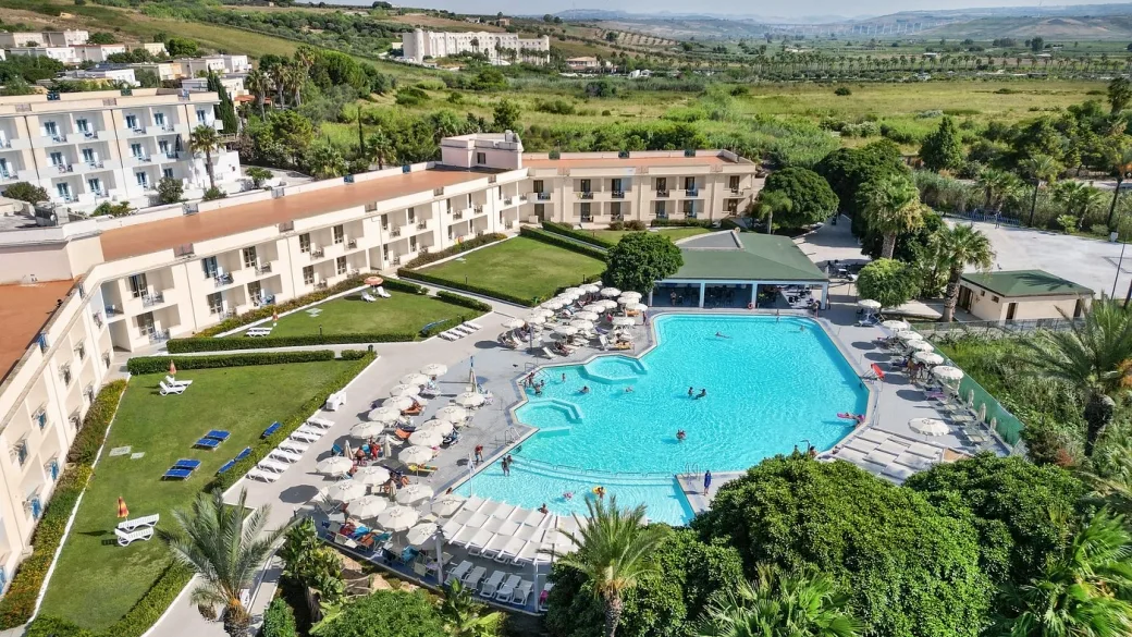 Vista aerea del resort con grande piscina, ombrelloni bianchi, giardino e colline verdi sullo sfondo