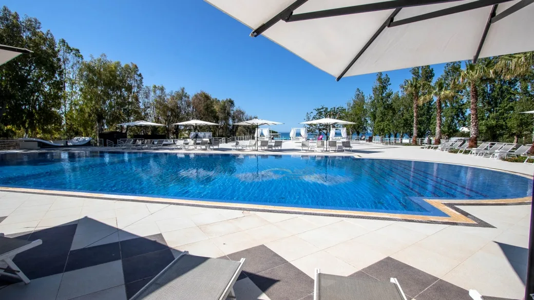 Ampia piscina esterna con lettini, ombrelloni bianchi e vista sul mare in giornata soleggiata