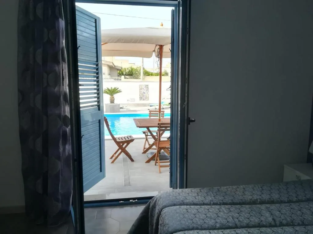 Vista dalla camera attraverso porta con persiane blu verso piscina e terrazza con tavolo