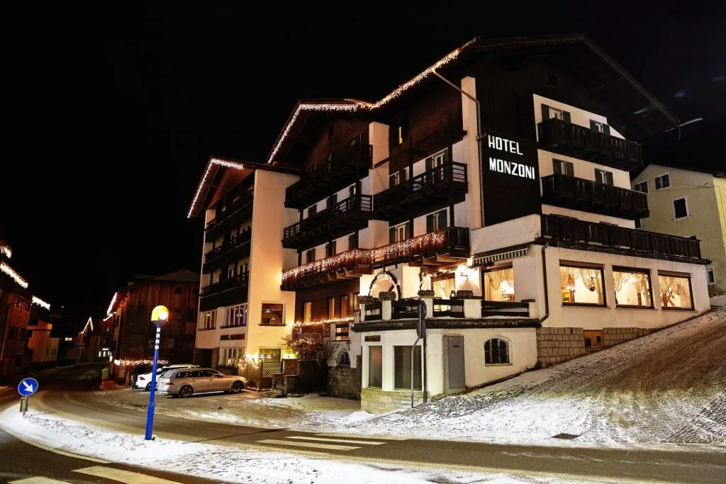 Facciata illuminata dell'Hotel Monzoni di notte con luci natalizie e strada innevata