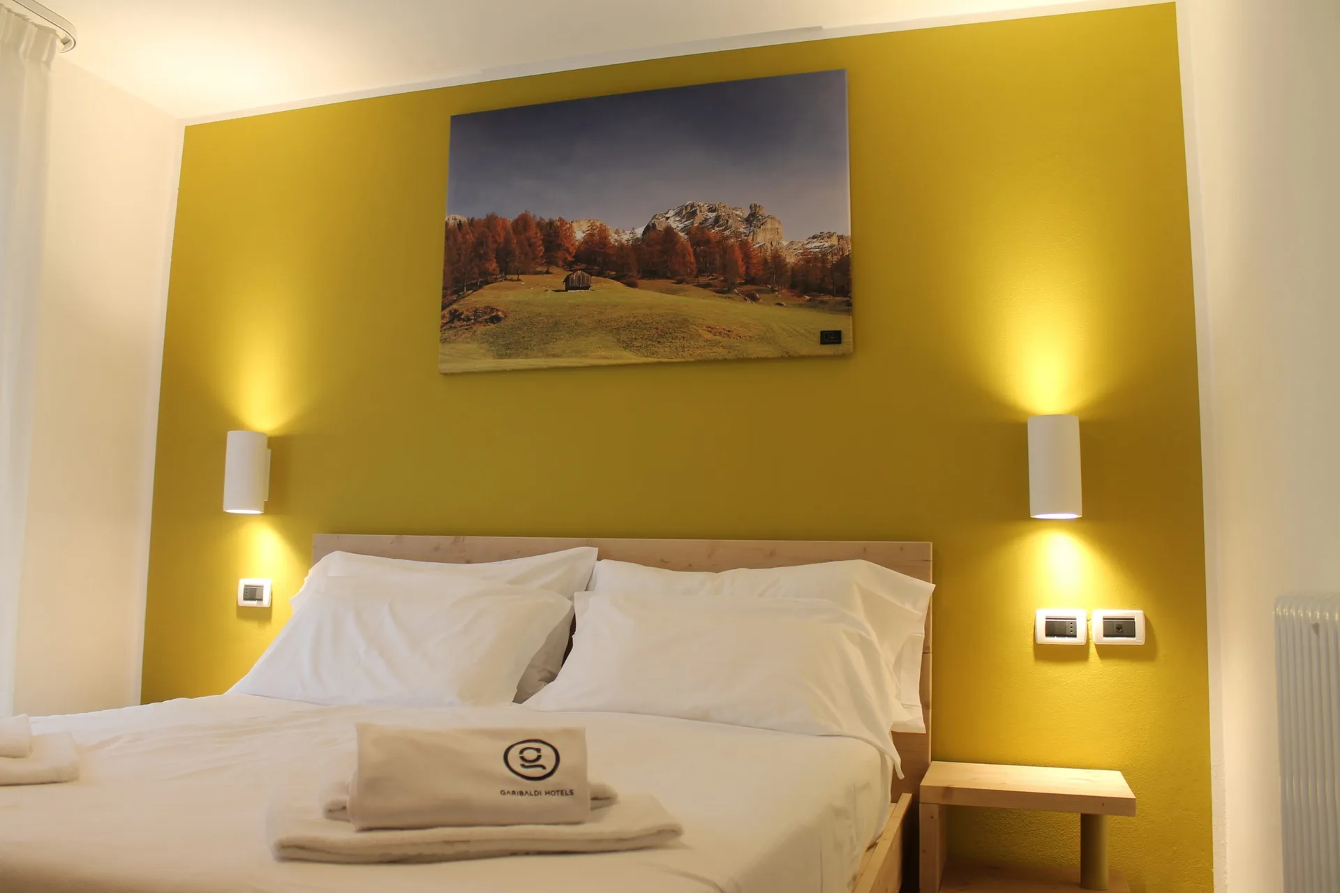 Camera matrimoniale con parete gialla, letto in legno e quadro con paesaggio autunnale dolomitico