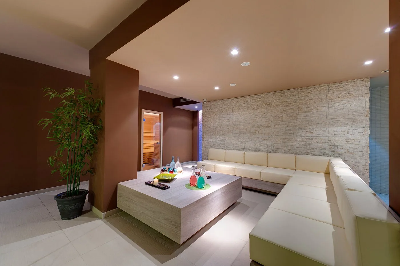 Area relax della spa con divani angolari, parete in pietra chiara e pianta di bambù