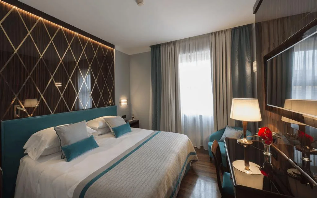 Camera elegante con testiera trapuntata a rombi, letto matrimoniale con dettagli turchesi, poltrona blu e illuminazione soffusa