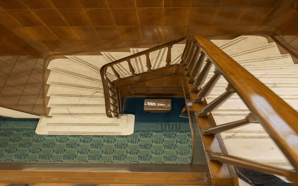 Scala a chiocciola con gradini in marmo bianco, corrimano in legno e tappeto verde vista dall'alto