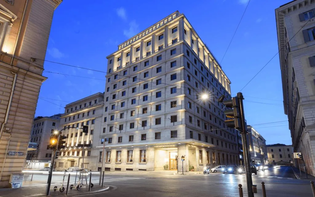 Facciata dell'hotel in stile razionalista illuminata al crepuscolo, situato all'angolo di una via cittadina