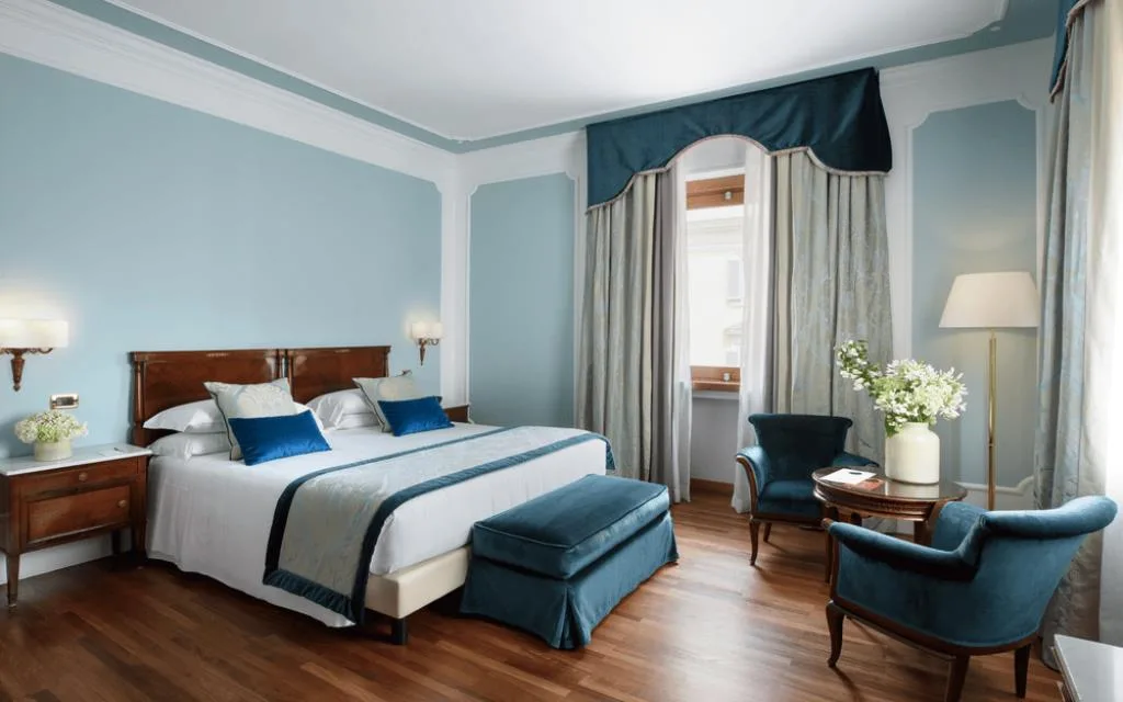 Camera raffinata con pareti azzurre, letto matrimoniale con testiera in legno, cuscini blu, poltrone e pouf in velluto e pavimento in parquet