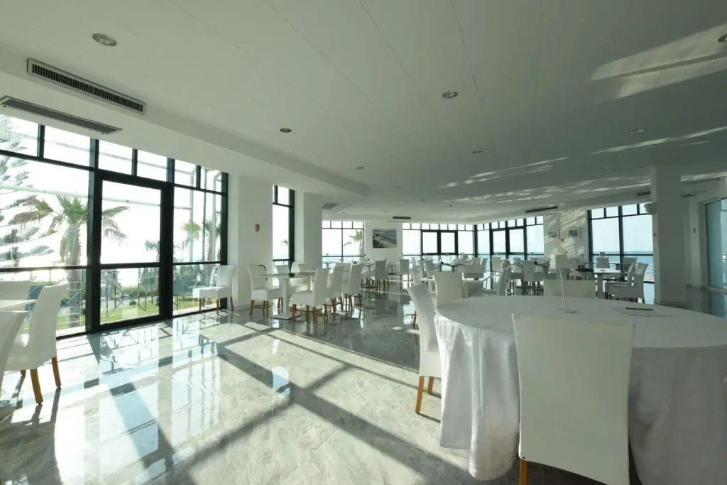 Sala ristorante luminosa con tavoli rotondi bianchi e ampie vetrate sul mare