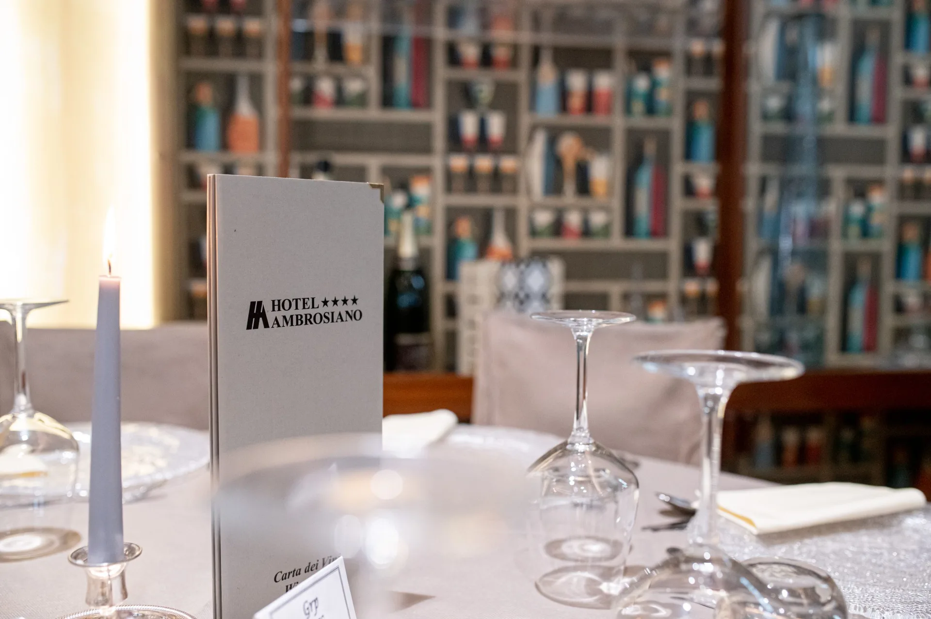 Tavolo apparecchiato con menu Hotel Ambrosiano, candela e cantina di vini sullo sfondo