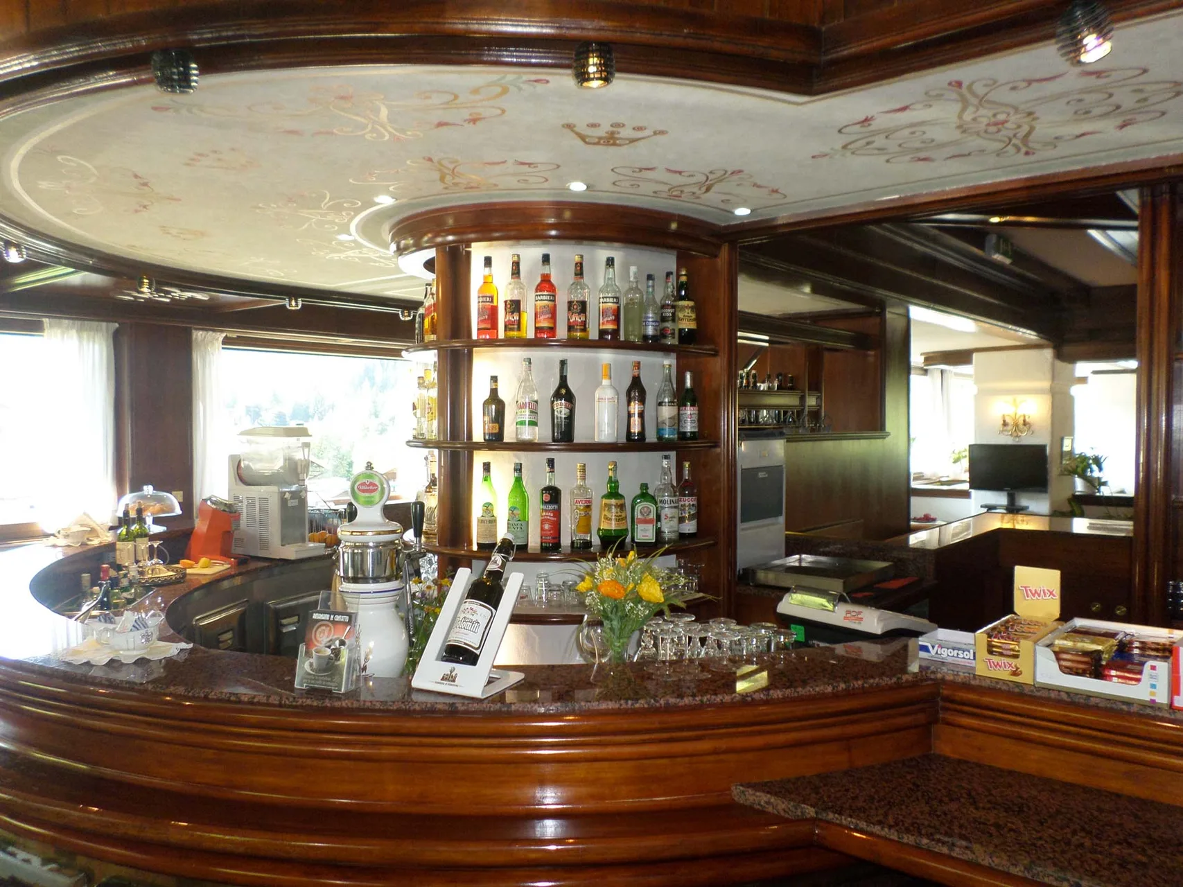 Bancone bar in legno curvo con bottiglie di liquori e soffitto decorato