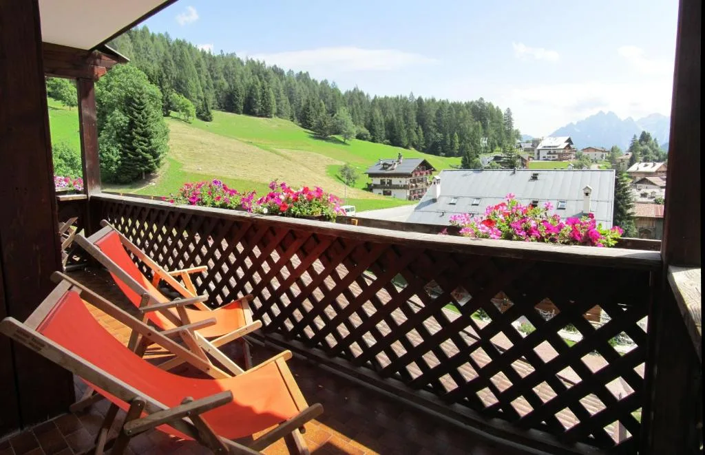 Balcone in legno con sedie a sdraio arancioni e fiori, vista su prati e Dolomiti