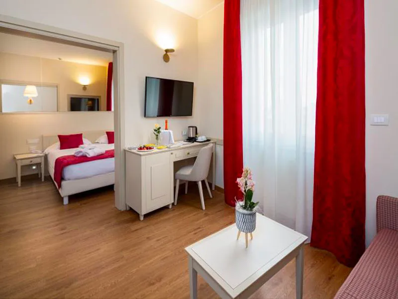Suite con zona giorno, tende rosse, scrivania bianca e camera da letto comunicante