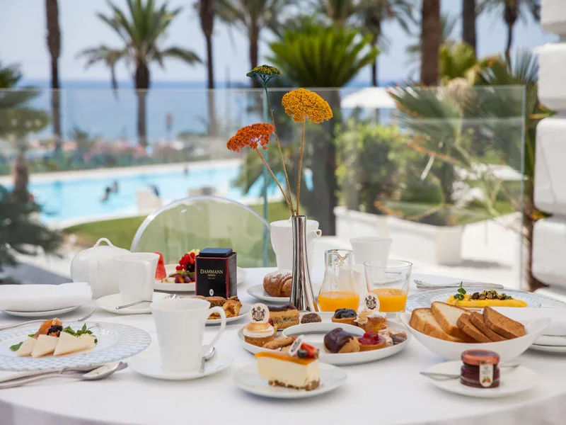 Colazione servita su tavolo con dolci, pane e frutta e vista su piscina e mare tra le palme